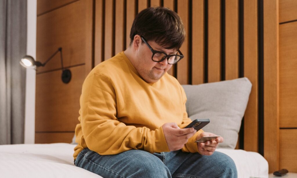 A man sitting on a bed looking at a phone and holding a bank card